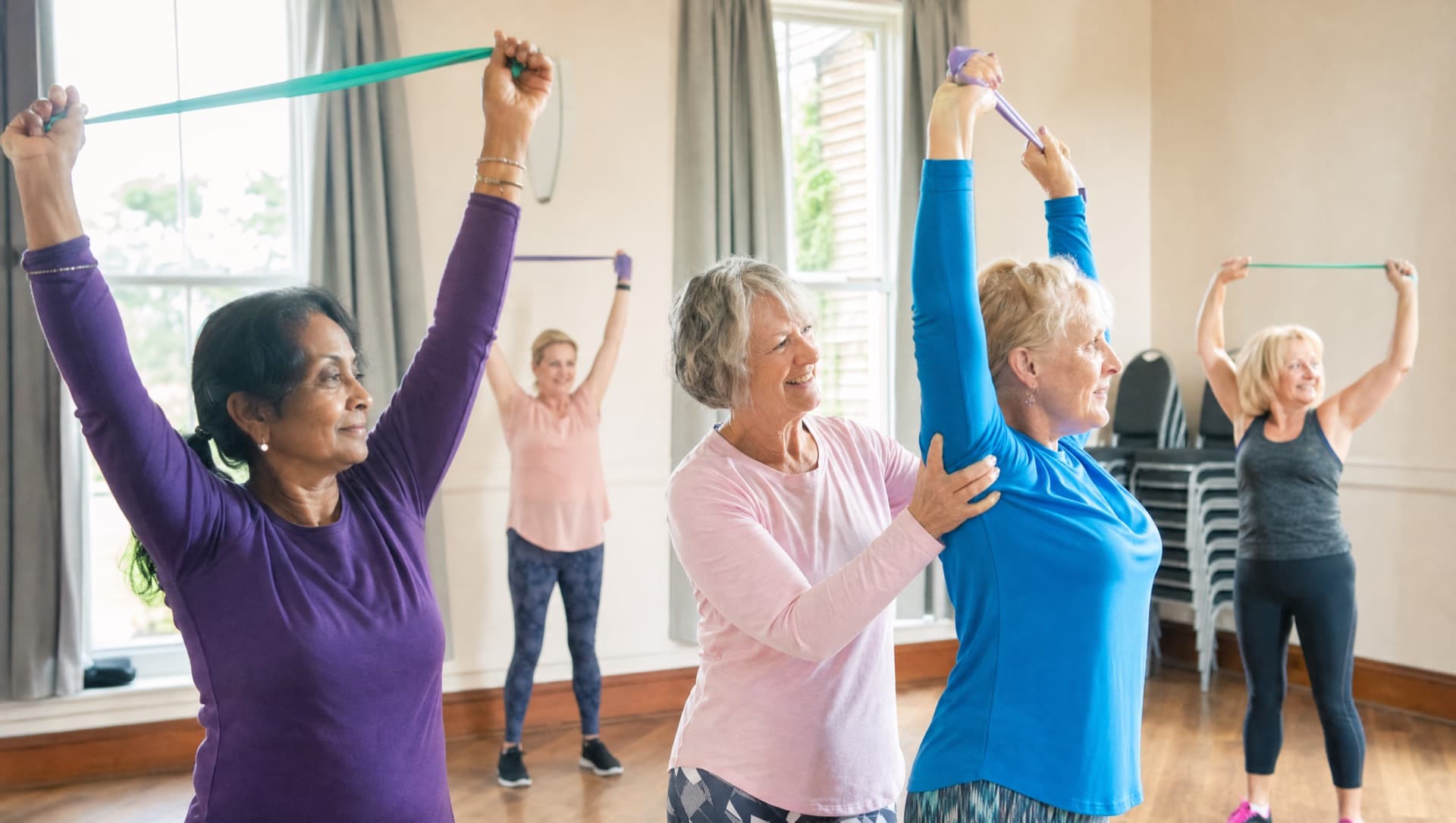 A small In the Pink rehabilitation class: women in their fifties and sixties stretching resistance bands in a bright village hall while a teacher guides one participant